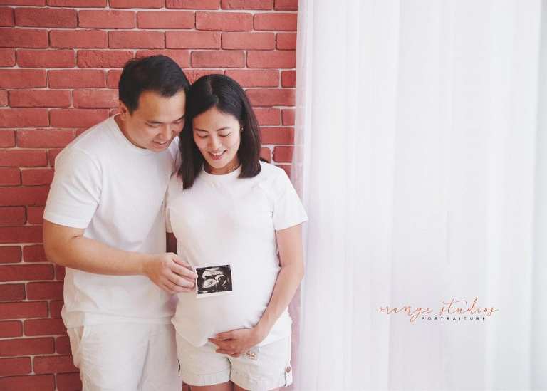 maternity portraits with husband against brick wall holding ultrasound scan in singapore studio with natural light