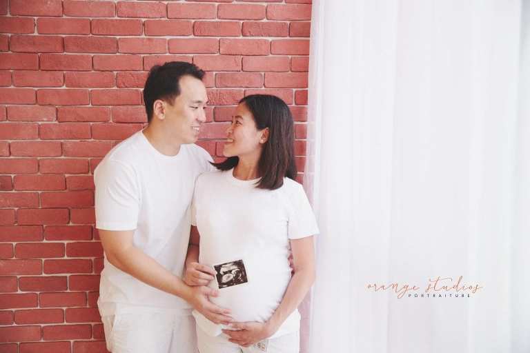 maternity portraits with husband against brick wall holding ultrasound scan in singapore studio with natural light
