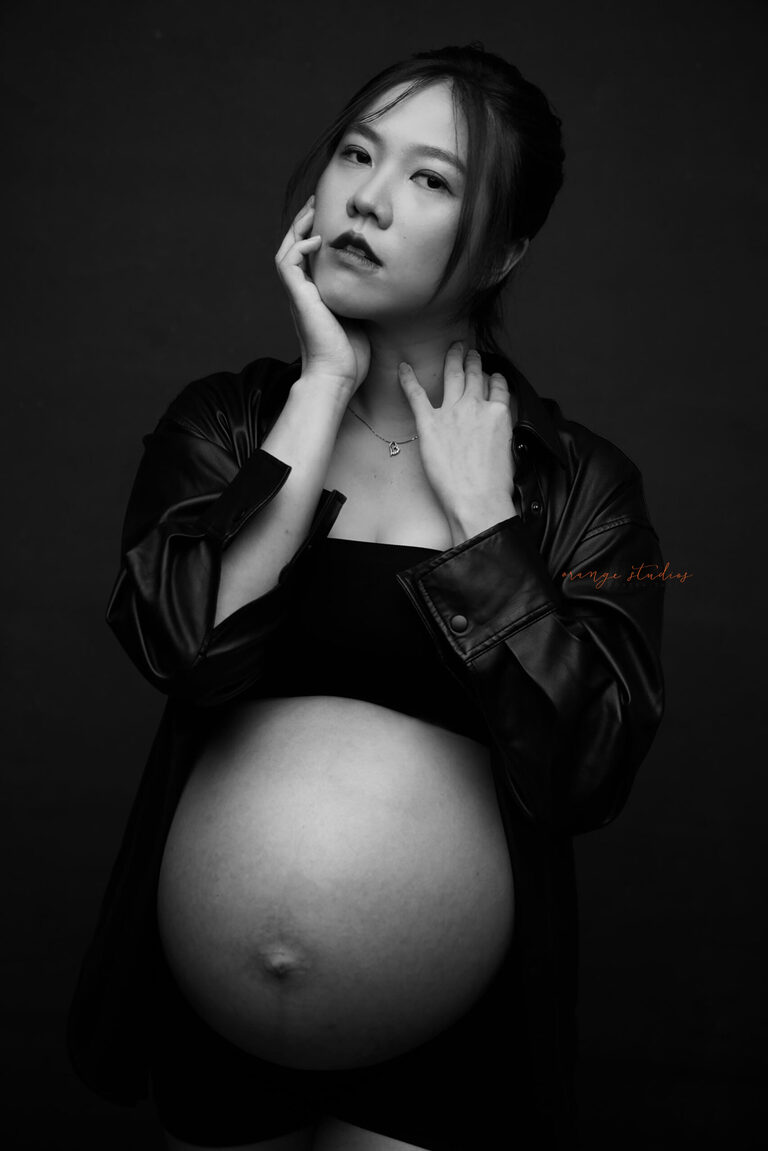 Dramatic black-and-white maternity portrait of a mum-to-be posing with hands on face, styled in a leather jacket — Orange Studios Singapore.