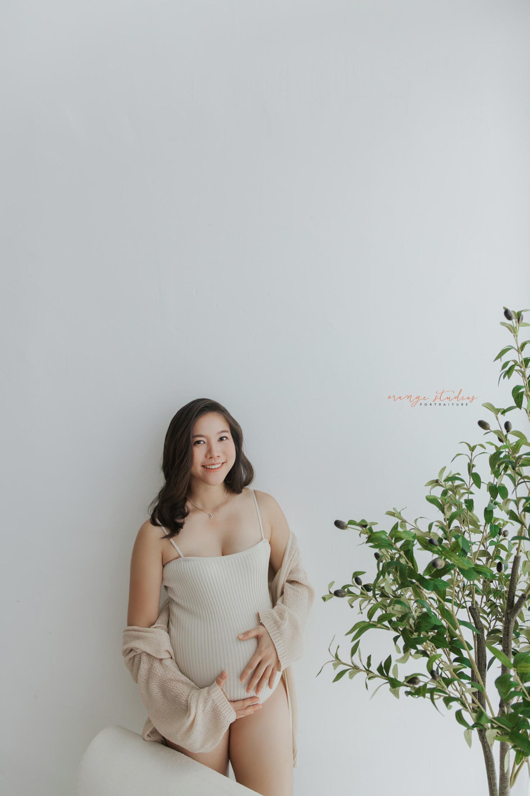 Bright and airy maternity portrait of a smiling mum-to-be in a cream outfit, styled with greenery for a natural look — Orange Studios Singapore.