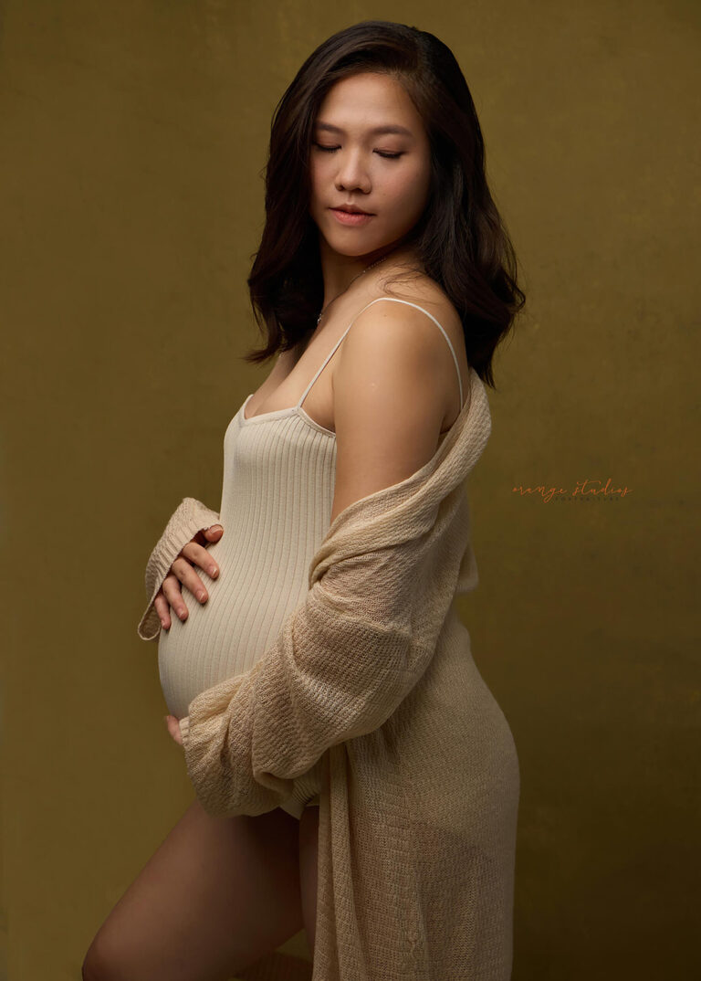 Studio maternity portrait of an expecting mum in a neutral beige outfit, softly lit against a warm golden background — Orange Studios Singapore.